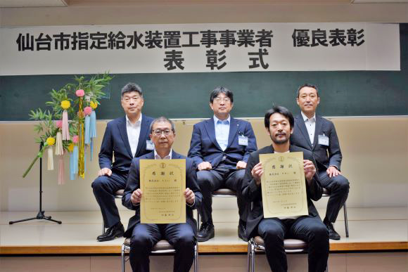 「仙台市指定給水装置工事事業者 優良表彰 表彰式」と書かれた横断幕の下で、表彰状を手にした2名の男性が前列に座り、3名の男性が後列に座っている記念写真