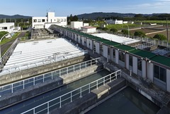 手前にコンクリート製のろ過池や水路が並び、その奥に白い管理棟、周囲には田畑や山々が広がるのどかな風景の中にある中原浄水場の写真