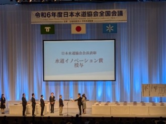 「令和6年度日本水道協会全国会議」の会場において、大型スクリーンに「水道イノベーション賞授与」と映し出される中、ステージ上で表彰状の授与が行われている様子を遠景から捉えた表彰シーンの写真