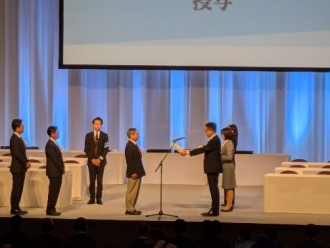 ステージ上においてプレゼンターから受賞者へ表彰状が手渡される瞬間を、周囲に並ぶ関係者とともに捉えた授賞式の写真