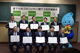 「青下の杜プロジェクトに関する協定締結式」と書かれた看板の前で、お揃いの書類を手にしたスーツ姿の男性10人と、右端に立つ水色で雫のような形をした水道局キャラクターウォッターくんが並んで記念撮影をしている集合写真