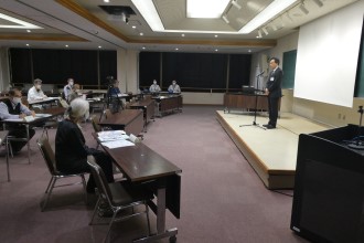 会議室で、前方のステージに立つスーツ姿の男性がマイクを通して話をしており、机に資料を広げて座る数名の参加者がその様子を静かに聞いている写真