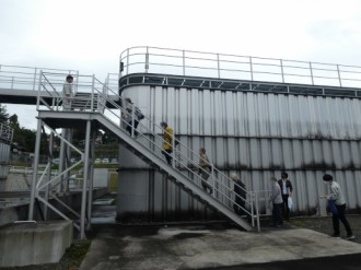 屋外にある巨大な金属製のタンクのような施設で、手すりの付いた外階段を数人の人々が一列になって上り、上部の通路へ向かって移動している様子を捉えた、荒巻配水所見学の写真