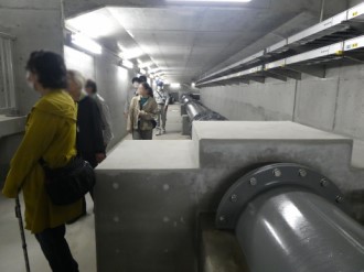 コンクリート壁に囲まれた通路のような場所で、太い金属製の配管が奥へと続き、その傍らを通路に沿って歩いたり立ち止まったりして施設内部を見学している数人の男女の写真