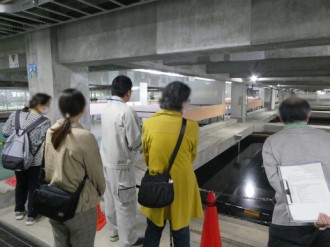 コンクリートの柱や梁が露出した屋内施設にて、作業服を着た案内員の後ろに並んだ数人の男女が、手前にある黒い水面が広がる貯水槽のような設備を熱心に見つめている、国見浄水場の見学の様子を背後から捉えた写真