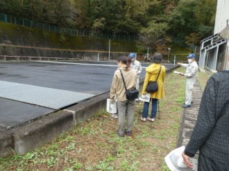 山際にある広い平坦な屋外施設で、作業服と青い帽子を着用した案内員が、資料を手に持った見学者の男女に向かって広大な敷地を指差しながら説明を行っている、上追沢沈砂池見学の写真