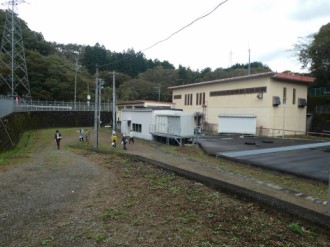 山々に囲まれた広い敷地内に、クリーム色の外壁を持つ施設と複数の小さな建物が並び、その建物の周辺や未舗装の広場を複数の見学者たちが散策したり移動したりしている様子を遠目から捉えた施設全景の写真