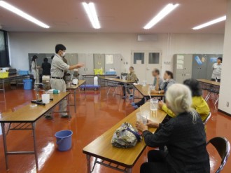 室内で、作業服姿の男性が長机を前にして身振り手振りを交えながら説明を行い、それを見守るように複数の男女がテーブルを囲んで座っている、実験器具や青いバケツが置かれた水質検査センターでの写真