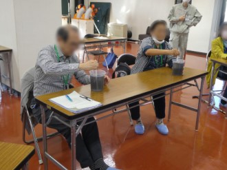 室内で、長机に座った男女がそれぞれ透明な容器に入った黒っぽい液体を棒のようなもので熱心にかき混ぜており、その様子を近くで作業服姿の人物が見守っている写真