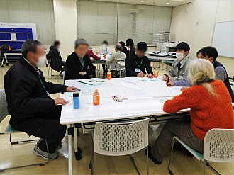 会議室のような場所で、男女数名が大きな白いテーブルを囲み、資料や筆記用具を広げながら真剣な様子で話し合いをしている様子の写真