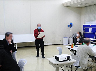 会議室のような場所の前方に、赤い服を着た男性が書類を手に持ちながら立ち、テーブルを囲んで座っている複数の男女に向かって意見を発表している様子の写真