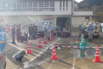 屋外のグラウンドで、ヘルメットを被ったスタッフが見守る中、カラーコーンで仕切られたエリアに置かれたホースから勢いよく水が噴き出す様子を人々が見学している写真