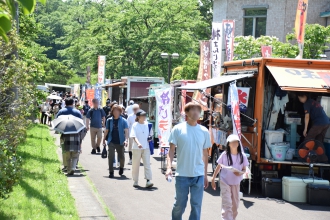 多くのキッチンカーやのぼり旗が並ぶ屋外通路で、家族連れなどの来場者が歩きながら買い物を楽しんでいる写真