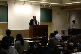 照明が落とされた会議室で、前方の壇上に立つ男性がマイクに向かって話し、手前に座る多くの出席者がその話を聞いている写真