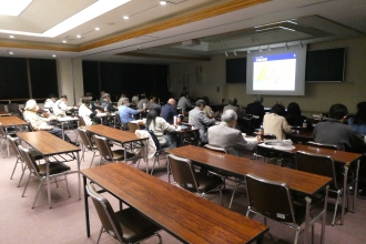 照明が落とされた会議室で、前方のスクリーンに映し出された資料を見ながら、多くの出席者が机に座って説明を聞いている写真