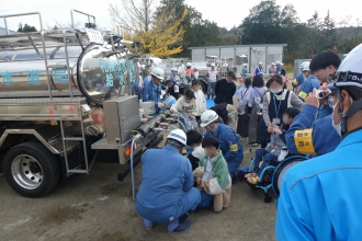 「札幌市水道局 給水車」と書かれた給水車の後ろで、ヘルメットを被ったスタッフのサポートを受けながら、子どもたちが蛇口から袋に水を入れる体験をしている写真