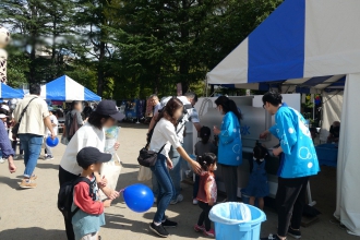 屋外のイベント会場で、青い法被を着たスタッフが並んでいる親子連れにコップを手渡し、給水タンクから注いだ水を試飲してもらっている写真