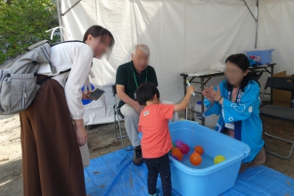 テントの下で、青い法被を着たスタッフや保護者に見守られながら、男の子が水色のタライに浮かんだカラフルなヨーヨー釣りに挑戦している写真