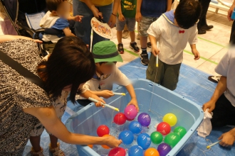 水色のタライに浮かんだカラフルなヨーヨーを、子どもたちが釣り針のついた棒を使って釣り上げようとしている写真