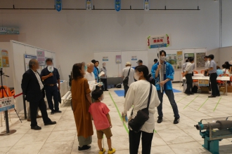 「最後尾」と書かれたプラカードを持つ青い法被姿のスタッフの周りに、展示パネルを見学したり順番を待ったりしている来場者が集まっている屋内イベント会場の写真