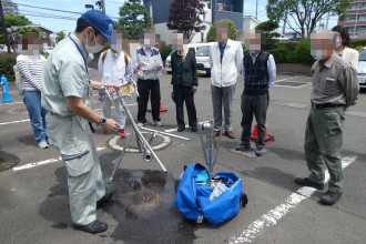 屋外の駐車場で、青い帽子と作業服を着用した男性がマンホールの上に三脚のような器具を設置しており、その周囲で男女数名が作業の様子を熱心に見守っている写真