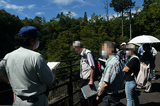 青空の下で生い茂る木々を背景に、青い帽子を被った作業服姿の男性が手元の資料を見ながら、手すり越しに景色を眺める男女数名に対して屋外で解説を行っている写真