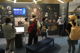 展示室内で、年表や資料が詳しく掲示された大きな壁面パネルを前に、スタッフの説明を熱心に聞き入る数名の見学者たちの後ろ姿を捉えた施設見学の様子写真