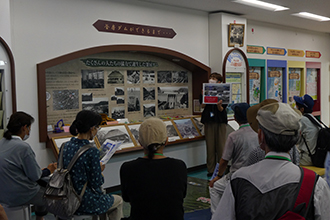 屋内展示室に設けられた、ダムの歴史に関する写真やパネルの壁面掲示の前で、案内スタッフの説明を熱心に聞く男女数名の参加者の後ろ姿を捉えた写真