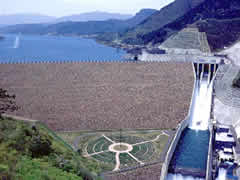 山々に囲まれた青い貯水池を背景に、広大で緩やかな傾斜を持つ茶褐色のロックフィル堤体と、右側に設置された滑り台のような形状の巨大なコンクリート製放水路から水が流れ落ちる様子を高い視点から見渡すように撮影した七ヶ宿ダムの写真