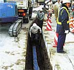 道路の舗装を剥がして深く掘削された溝の中で、ヘルメットを被った作業員が青い配管の敷設状況を確認しており、地上では重機や交通整理のスタッフが安全を確保しながら水道管の工事を進めている水道管工事現場の写真
