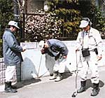 屋外の路上にて、作業着とヘルメットを着用した3人の作業員が、音聴棒などの器具を地面に当てたりヘッドホンで音を確認したりして、地下に埋設された水道管の漏水箇所を特定しようとしている様子の写真