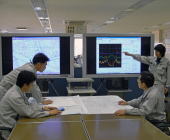 作業着を着用した4人の人物が、室内の大きなモニターに表示されたグラフや図面を指し示しながら、手元の資料と照らし合わせて分析を行っている写真