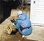 屋外の敷地内にて、青い作業服と帽子を着用した作業員が地面にある水道メーターの蓋を開け、手に持ったハンディターミナル端末を操作して、検針作業を行っている後ろ姿を捉えた写真
