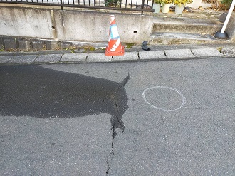 アスファルトの路面に生じた亀裂から水が染み出し、周囲が濡れている様子とともに、漏水箇所の目印として路面に描かれた白い丸印や、注意喚起のために置かれたカラーコーンを捉えた、地下の配管損傷による漏水現場の写真