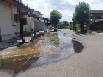 住宅街の道路において、路面から湧き出した水が、アスファルトの上を川のように長く流れ広がっている様子を捉えた、地下の配水管の破損による大規模な漏水現場の写真