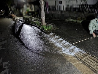 夜間の住宅街において、路面の亀裂から勢いよく水が噴き出し、車道に広がっている様子を捉えた、夜の漏水現場の写真