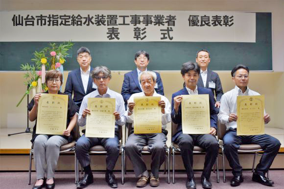 「仙台市指定給水装置工事事業者 優良表彰 表彰式」と書かれた横断幕の下で、表彰状を手にした5名の男女が前列に座り、3名の男性が後列に座っている記念写真