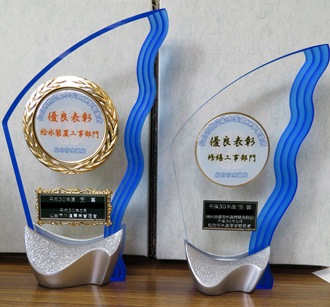「給水装置工事部門」と「修繕工事部門」と記された、波のような青いラインが特徴的な、優良表彰記念品のクリスタルトロフィーが2つ並んだ写真