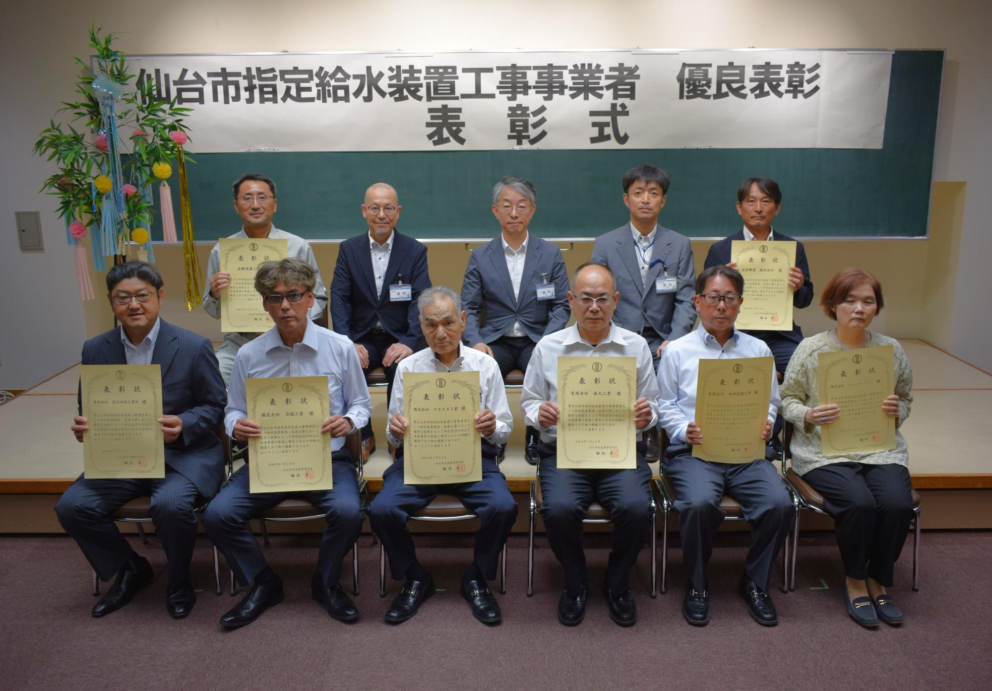「仙台市指定給水装置工事事業者 優良表彰 表彰式」と書かれた横断幕の下で、表彰状を手にした8名の男女が前列と後列に座り、3名の男性が後列に座っている記念写真
