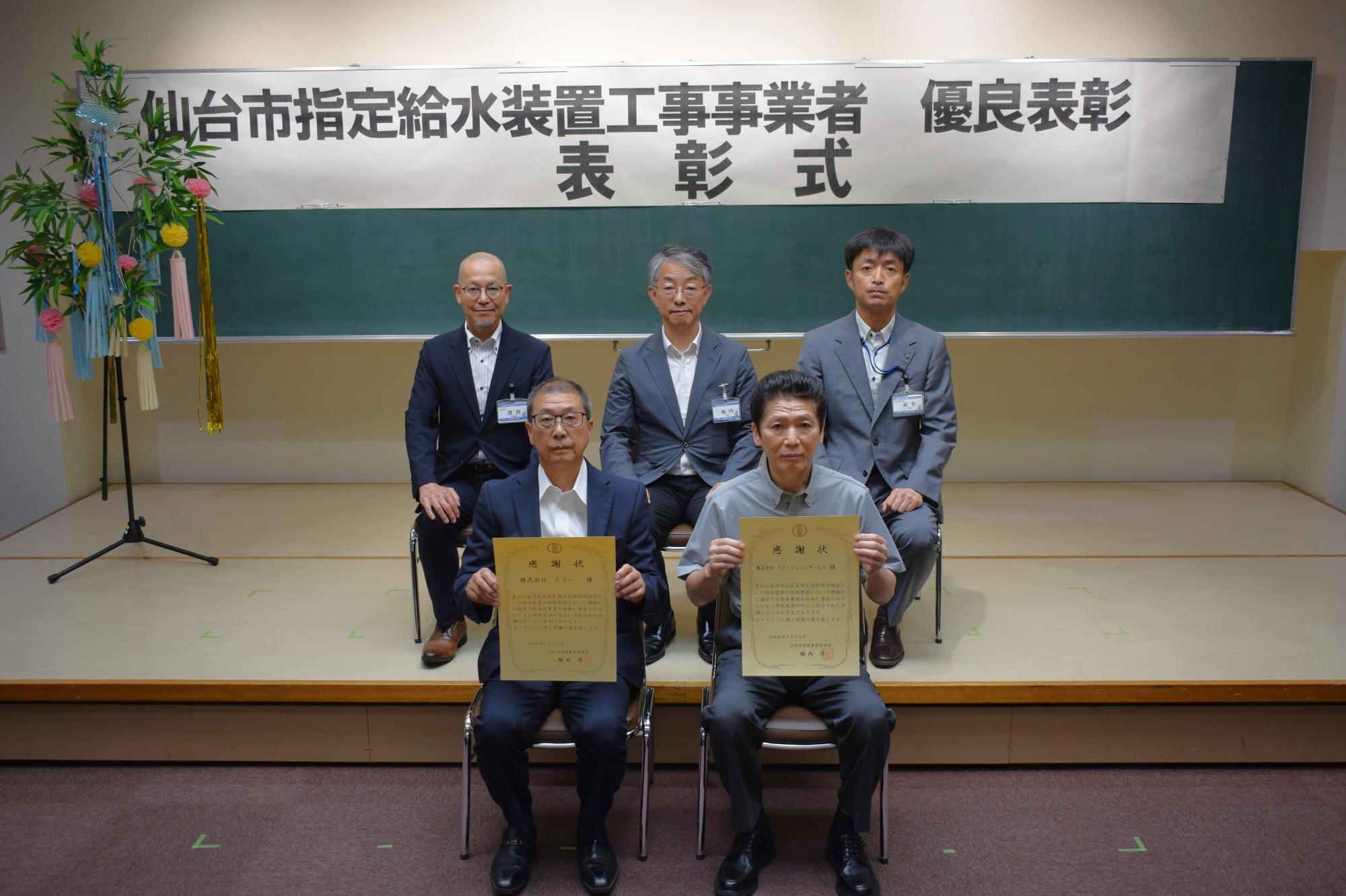 「仙台市指定給水装置工事事業者 優良表彰 表彰式」と書かれた横断幕の下で、表彰状を手にした2名の男性が前列に座り、3名の男性が後列に座っている記念写真