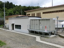 浄水場の白い平屋建ての建物の横に設置された、ボックス型の上追沢沈砂池小水力発電設備建屋の外観写真
