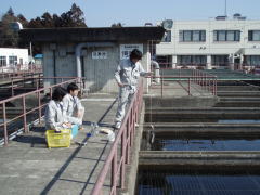 作業服を着た3人の職員がコンクリートの水槽脇の通路で水を採取をしている様子の写真
