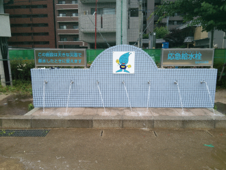 水色の文字で「応急給水栓」と書かれたシルバーの看板があり、中央に水道局キャラクターのウォッターくんが描かれたタイルの壁に設置された複数の蛇口から水が出ている、屋外の災害時給水栓（水飲み場型）の写真