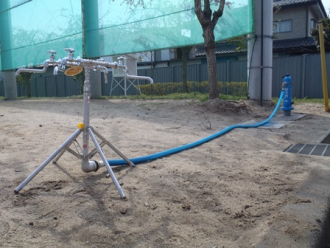 砂地に設置された青い給水栓から青いホースが伸び、三脚で自立する複数の蛇口を備えた仮設給水装置に接続されている災害時給水栓（地上型）開設時の写真