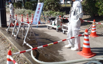 三角コーンとポールで区切られた区域に「臨時給水所」の看板と仮設蛇口スタンドが設置され、職員が立つ非常用飲料水貯水槽の給水所開設状況の写真