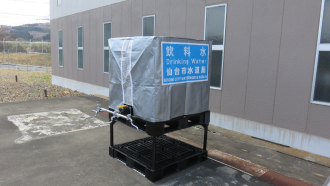 建物脇の屋外で、シルバーのカバーで覆われ「飲料水 仙台市水道局」という青いパネルが貼られた蛇口付きの四角い水槽が、黒い架台の上に設置されている、組立後の仮設水槽の写真