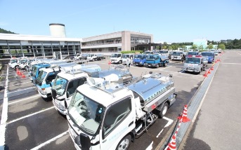 銀色のタンクを積んだ多数の給水車や、作業用トラックが、建物前の広い駐車場に整列して停車している様子の写真