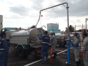 駐車場に停車している給水車のタンクに、複数の作業員が給水設備を用いて注水作業を行っている様子を捉えた写真