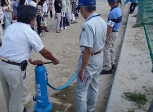 屋外に集まった人々の前で、職員の監督の下、青い給水栓を男性が操作している災害時給水栓の設営訓練の写真