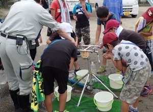 屋外に開設された災害時給水栓の蛇口から複数の子どもたちが水を飲んでいる様子の写真